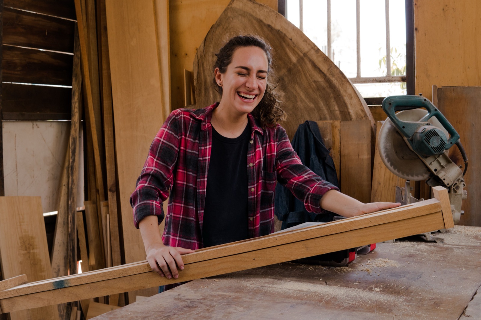 Lachende vrouw in een houtwerkplaats aan het werk met een plank, als beeld van zingeving in je werk