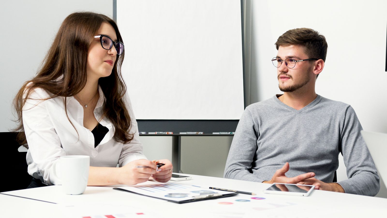 Twee collega's aan een vergadertafel met documenten, de een stelt een vraag en de ander luistert aandachtig