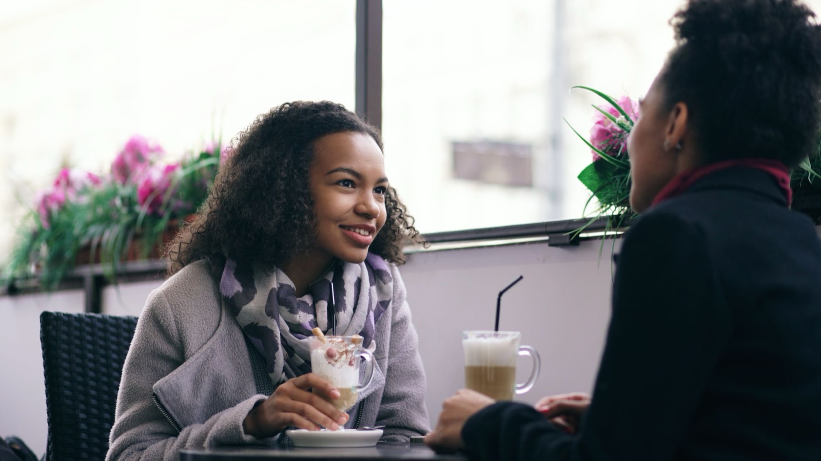 Twee vrouwen in gesprek aan een cafétafel; de een luistert aandachtig naar feedback over hoe die overkomt
